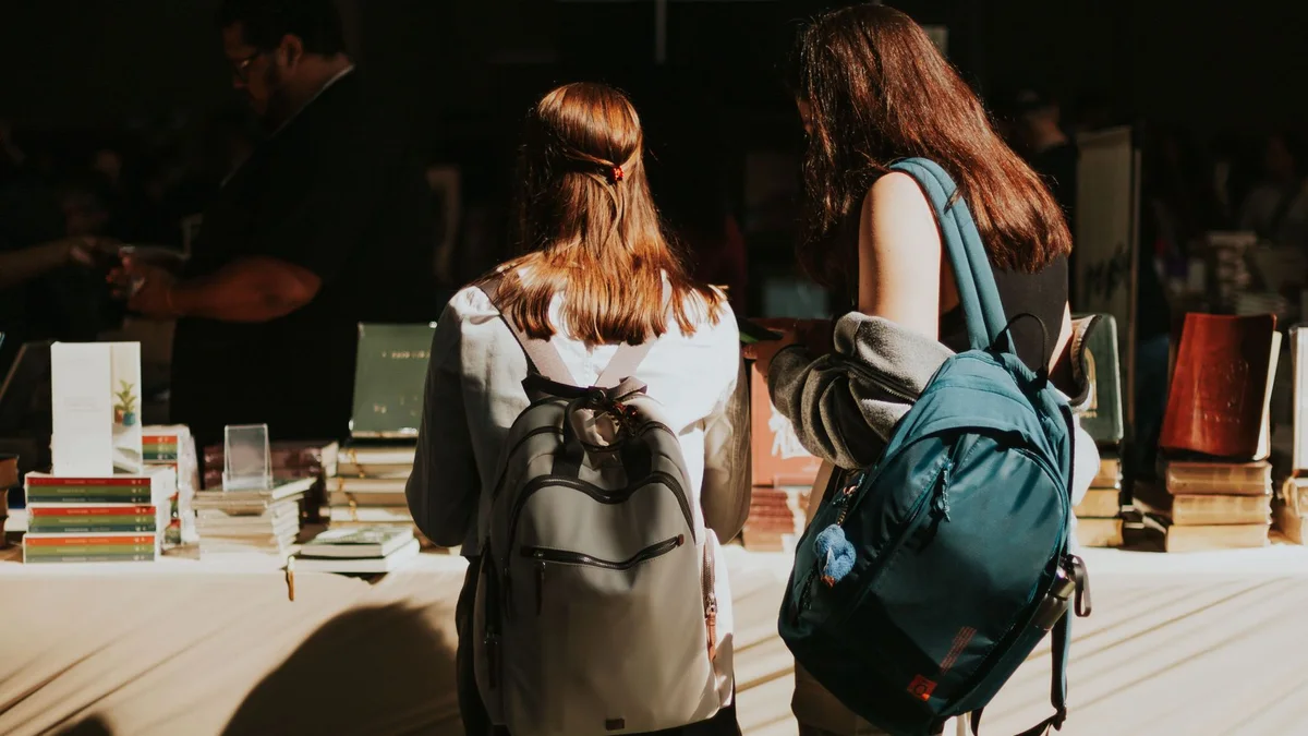 Duas estudantes com mochilas observando livros expostos em uma feira